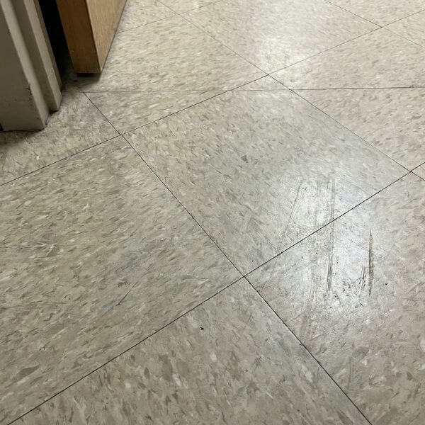 A close-up view of scuffs and scratches on worn commercial vinyl floor tiles showing wear and tear.