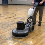 A service technician operates a commercial stripping machine on a section of worn wooden gym flooring.