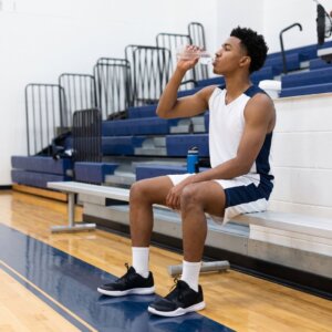 A student-athlete in a white jersey sits on the sidelines of a clean gymnasium floor, drinking water.