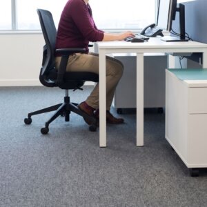 A corporate employee works at a workstation with grey carpeting in an open-plan office.