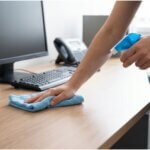 A person in an office uses a microfiber cloth and spray to clean a wooden desk surface near a computer monitor.