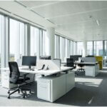 A wide, sunlit modern office workspace viewed through floor-to-ceiling windows, showing a clean, safe environment.